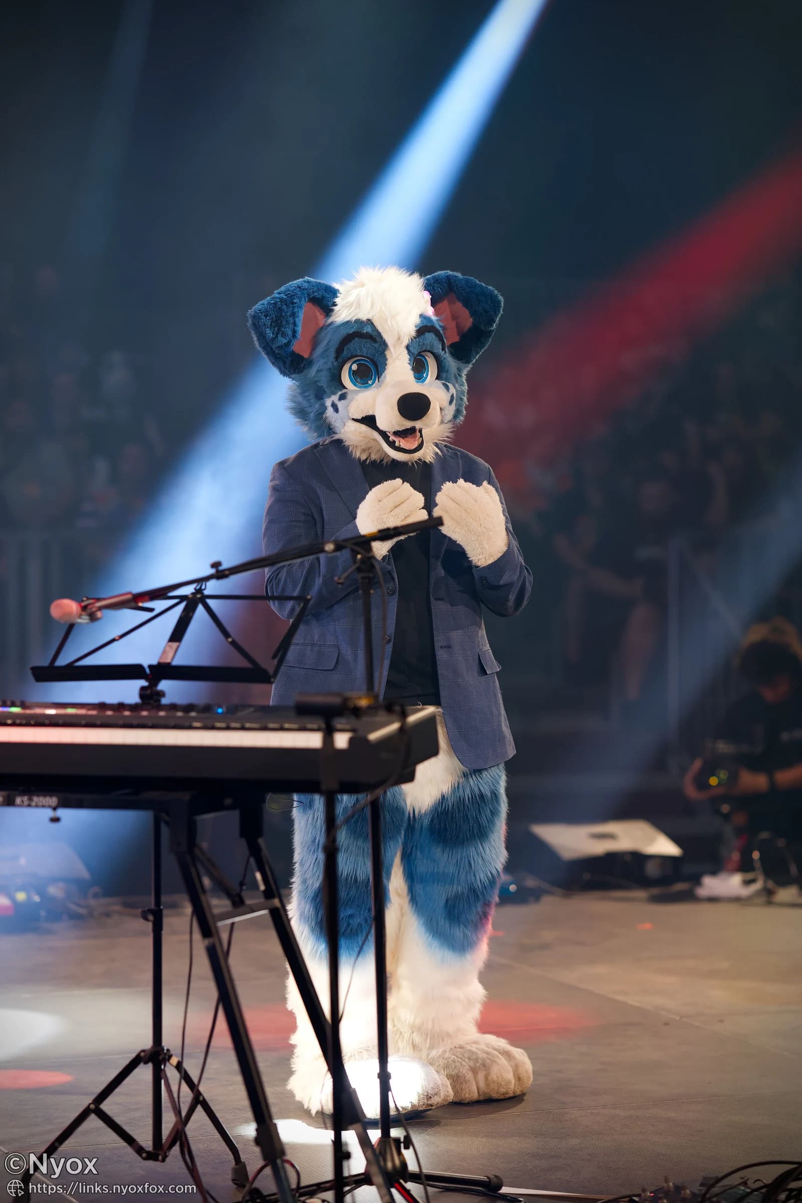Little Alpha Pup performing on stage in a blue and white fursuit and checkered blazer, standing at a keyboard under stage lights